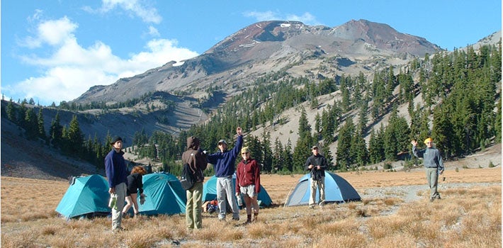Students Camping at Base of Mountain Several students stand outside their tents in a field of long, yellow grass, at the base of a mountain.