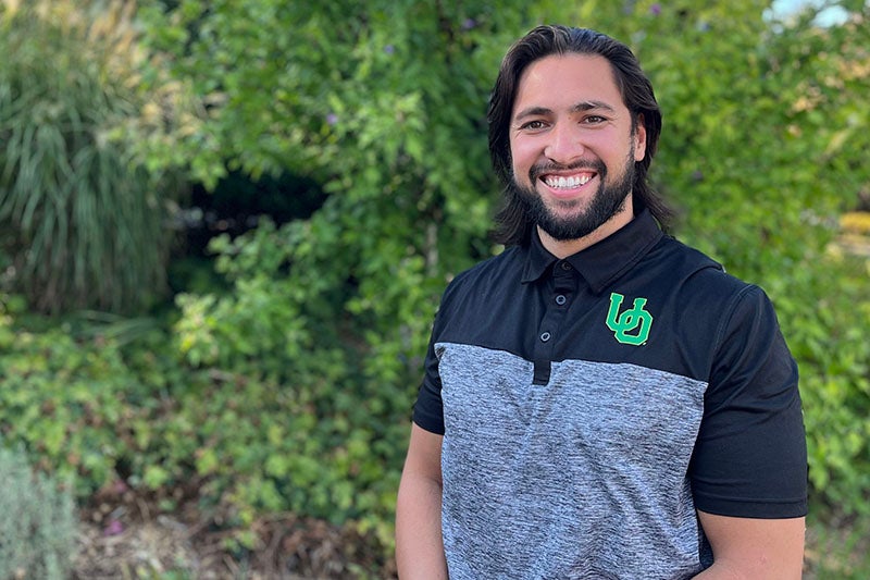 Profile photo of Caleb, wearing a black-over-grey UO-branded polo shirt, in front of trees and ivy