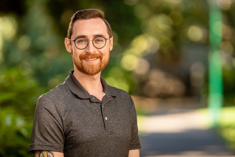 Profile of Kyle, standing in front of shrubbery, and a walkway leading to the background