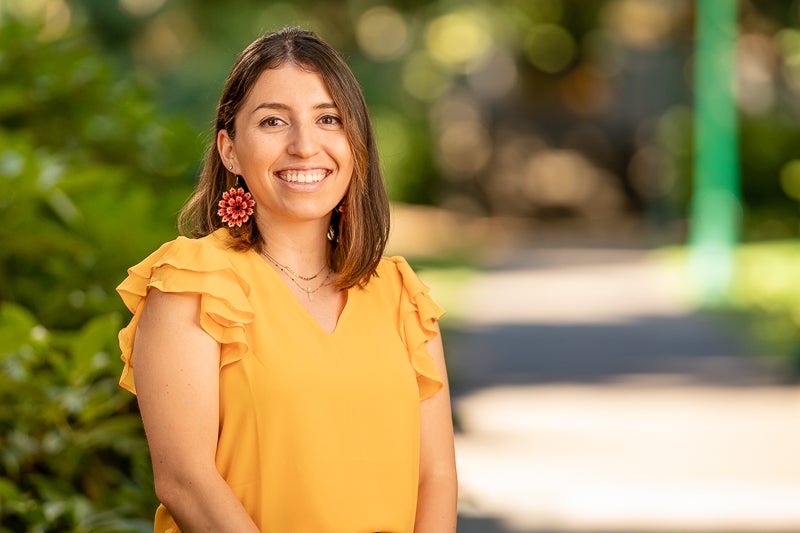 Yollixpa wearing a yellow-orange top, standing in front of shrubbery and a pathway blurred in the background