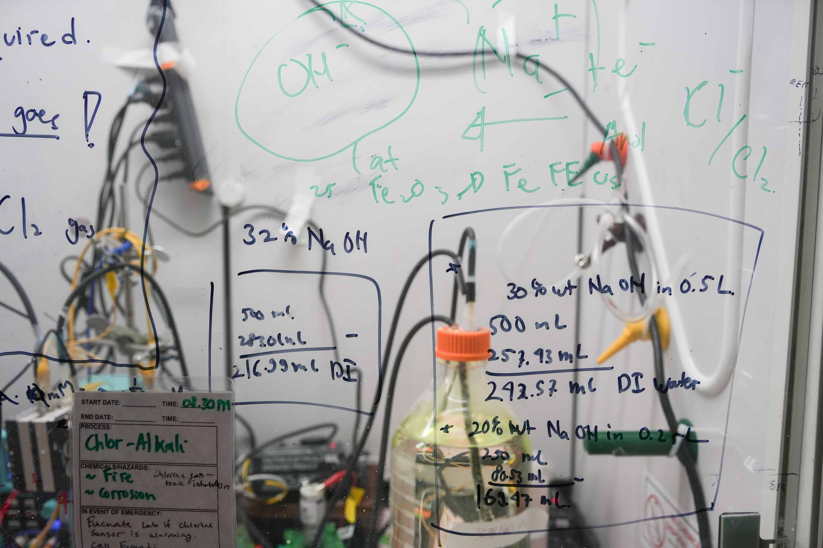 Materials science and technology laboratory bottles and cables are connected behind a clear surface covered with written scientific notations
