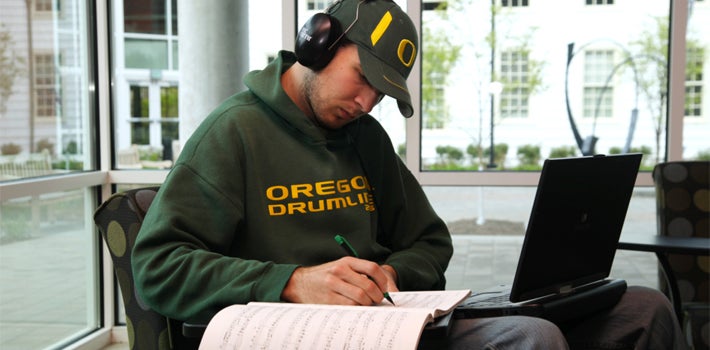 A man wearing an "Oregon Drummer" sweatshirt writes music while wearing headphones.